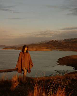 A woman in a brown wool poncho strides up an incline with a backdrop of hill chains and calm lake waters, seen at sunset.