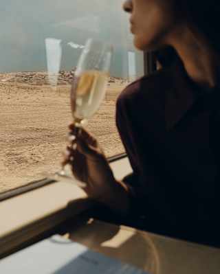 In soft-focus foreground, a seated woman holds her glass of sparkling wine at a window table as sand-hued plains pass by.