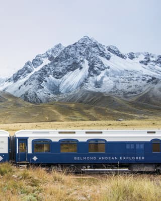 Two white and navy train carriages before snow coated Andean mountains