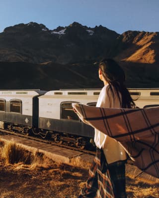 A Peruvian woman stands in the grass alongside the train track, watching the Andean Explorer pass, with the wind in her hair.
