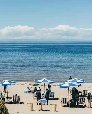 People relax on a sandy beach under blue and white parasols, facing a calm, expansive body of water with distant mountains and clouds on the horizon under a clear blue sky.