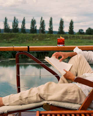 A man in casual clothes reclines in a chair, reading a book with a Negroni-style drink next to him on a deck's wooden rail.