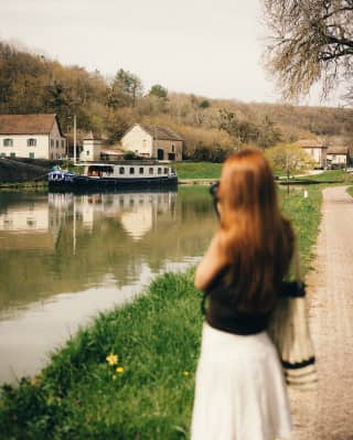 A woman stands on the bank of the canal, taking a photograph of the Fleur de Lys as it cruises by a cluster of white houses.