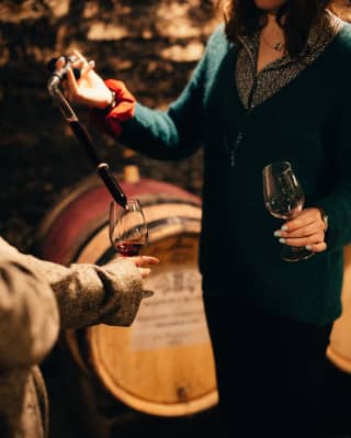 By a row of oak wine barrels in the cellar of a Burgundy vineyard, an expert pours red wine into a glass for a guest to try.