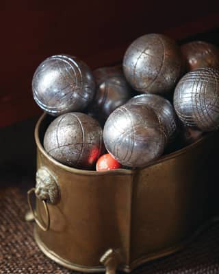 A brass bucket with a lion's head feature and claw feet, containing well-used, steel petanque balls and a red wooden jack.