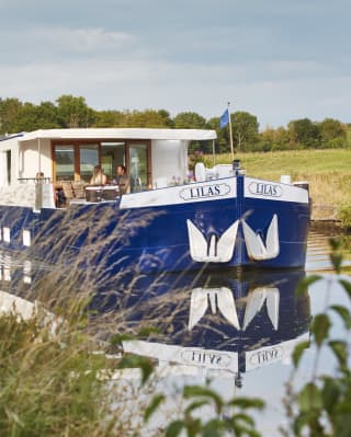 The Lilas cruising with two passengers on deck enjoying the sunshine and tranquillity, pictured through grasses on the bank.