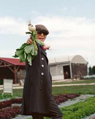 Chef Dominique Crenn, in dark glasses and a flat cap, stands in a vegetable farm, holding up a bunch of fresh-pulled turnips.