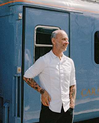 A man with tattoos wearing a white shirt stands and smiles in front of a blue train carriage on a sunny day.