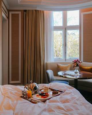 A silver cloche rests by a tray carrying cooked breakfast, coffee, orange juice and a pastry, set on bed in an elegant suite.