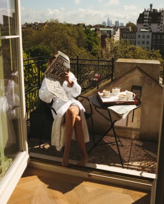 A female guest in a white bath robe reads a newspaper at a table set for morning coffee on a balcony with big views.