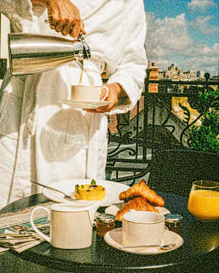 A person in a white dressing gown pours coffee into a cup on a balcony table set with pastries, a pudding, juice, and small jars, with a cityscape and blue sky in the background.