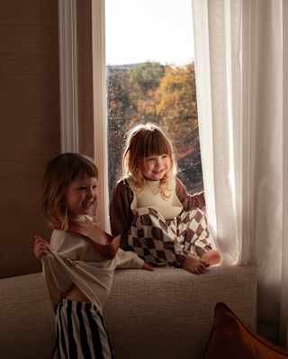 Two young children with shoulder-length hair sit and stand near a window with sheer white curtains, smiling and enjoying the warm sunlight streaming into the room. Trees with autumn foliage are visible outside.