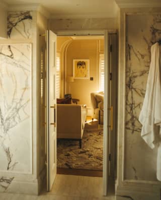 Looking from the marble-walled penthouse suite bathroom through the open door to the living area, bathed in a honeyed light.