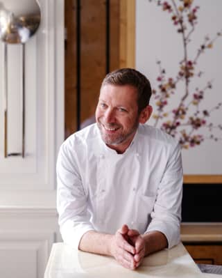 A portrait of renowned pastry chef, Benoit Blin, smiling to camera with arms resting on a marble counter with palms together.