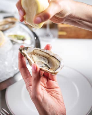 An open Porthilly oyster is held in a left hand, while the right hand squeezes half a lemon above it