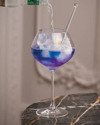 A bottle of tonic water is poured into a tall-stemmed globe glass containing a blue cocktail, glass stirrer and ice cubes