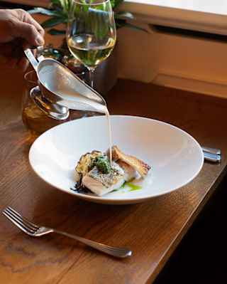 A waiter pours a cream sauce on to pieces of crispy-skinned grilled white fish with parsley in a close-up detail at The Lalee.