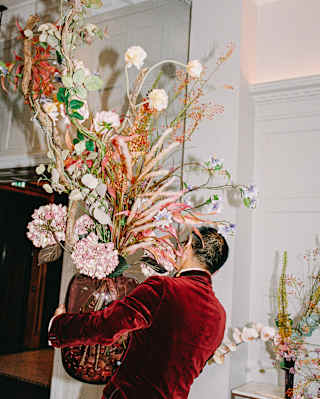 A man in a red velvet jacket carries a huge red glass vase of big blooms and tall stems in shades of pink, seen from behind.