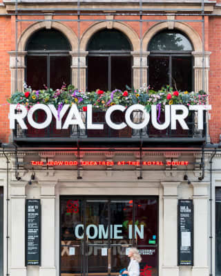 A woman walks beside the iconic front entrance of the Royal Court Theatre, its glass doors emblazoned with the words come in