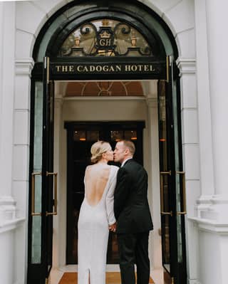 A blonde bride with a backless dress kisses her groom on the marble stoop of the elegant entrance to The Cadogan Hotel