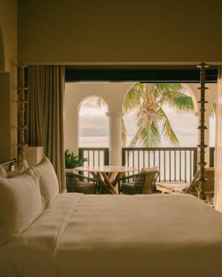 View over the white bed in an unlit room to the balcony with seating and sublime views of the sea, brushed by palm fronds.