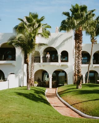 View from the garden to a white, Moroccan-style, two-level villa with arched colonnade balcony and terrace, flanked by palms.