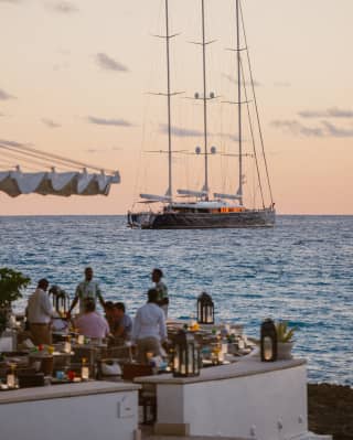 At sunset, guests mingle among candle-lit tables on Cip's terrace, enjoying views of the bay and a grand sailing yacht.