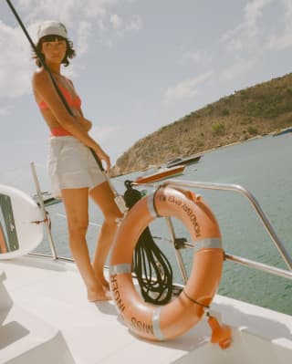 Enjoying a day on the water, a woman in an orange bikini top and white shorts stands at a boat's railings by a lifebuoy ring.