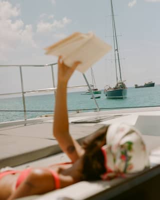 In soft-focus, a woman bathing on a boat deck shades her face with an open book as she reads with views of sailboats and sea.