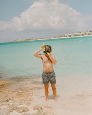 Facing the camera, a man in grey trunks adjusts his mask and snorkel as he stands in the shallows at the east end of bay.