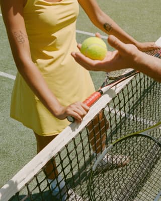 Close-up of a woman in a yellow dress and a man holding a tennis ball as they meet either side of the net with their rackets.