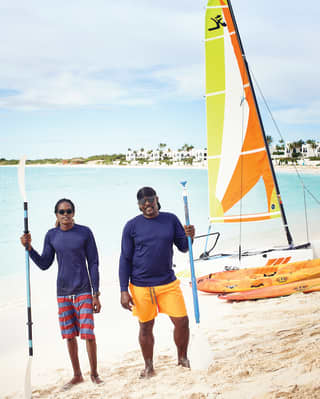 Two young men in navy long sleeve rash vests hold oars next to orange sea kayaks and a small sailboat on the sand