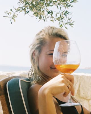 A young woman looks at the camera through the top of a glass of Aperol as she sits in the shade of an olive tree
