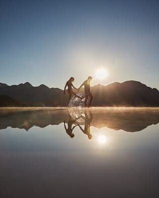A setting sun silhouettes the Lattari Mountains as a couple paddle hand in hand at the horizon edge of an infinity pool