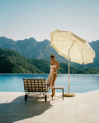 A woman in a white bikini stands with a towel next to her poolside lounger, beneath a parasol, with a mountainous backdrop.