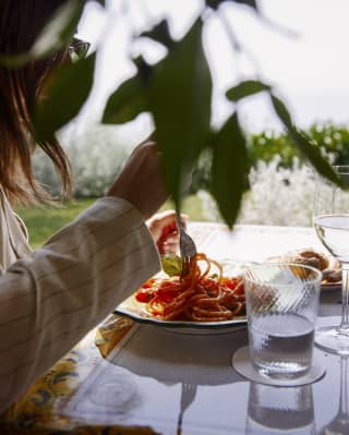A woman sits at a glass-topped table in a shady corner of a garden twirling marinara-drenched spaghetti with a fork