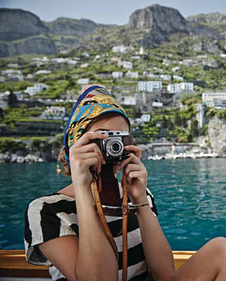 A woman in a headscarf and striped top takes a photo sitting on a small boat; behind her Ravello climbs the mountain sides