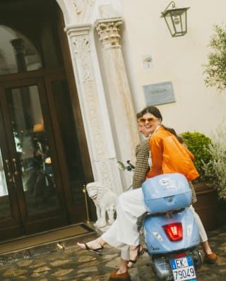 A smiling woman in an orange top and glasses sits side-saddle on the back a blue Vespa motorcycle behind a man in tweed.