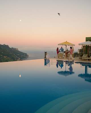 A reflection of the moon in a peach sky settles on the still water of the infinity pool as four guests enjoy poolside drinks.
