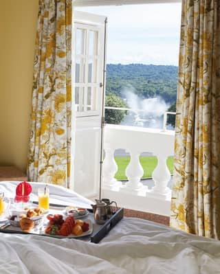 Breakfast tray including fruit juice, pastries and coffee on a hotel bed alongside open balcony doors with sunny rural view