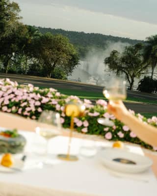 A hand raises a glass of wine at an al-fresco table in soft-focus foreground in an angled shot of the gardens and falls.