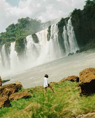 Angled image as a woman in shorts and white shirt walks across a grassy bank, gazing back at a stunning Iguassu waterfalls.