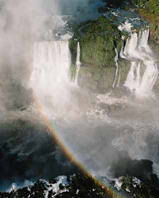 Aerial view of the Iguassu Falls in sun, cascading in streams over foliage-covered rocks, throwing up spray and a rainbow.