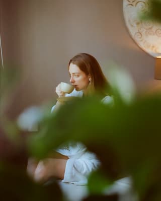 Seen from behind a plant, a woman with long, straight, brown hair sips tea in a white towelling robe in the peace of the spa.