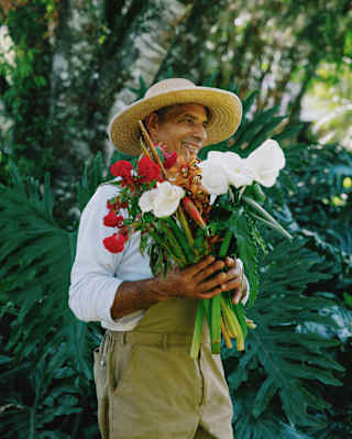 A smiling male gardener, in a hat, white shirt and khaki dungarees holds a large bunch of white amaryllis and red carnations.
