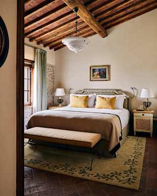 Beneath a pitched beam ceiling, a large bed in gold, wheat and taupe fabrics catches natural light from an adjacent window.