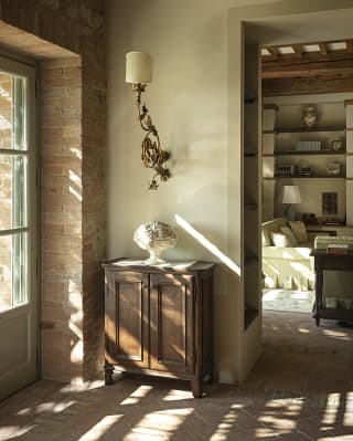 Kissed by light from a  window, a wood cabinet with a porcelain ornament sits against a wall under a filigree light fitting.