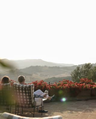 View from behind of two guests sitting on a terrace, relaxing with a glass of wine as they gaze out at the hazy Tuscan hills.