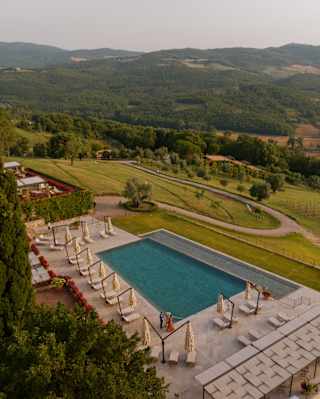 Loungers and cantilever parasols line three sides of the pool, with the infinity edge open to Tuscan hills, seen from above.