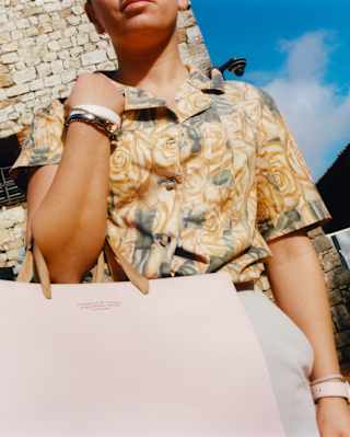 Low-angle shot of a woman in a floral blouse and bangles as she carries a pink shopping bag in the crook of her elbow.